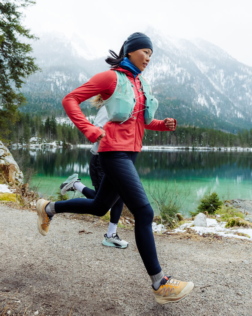 Woman running outdoors beside a lake wearing an insulated red jacket, mountains in the background.