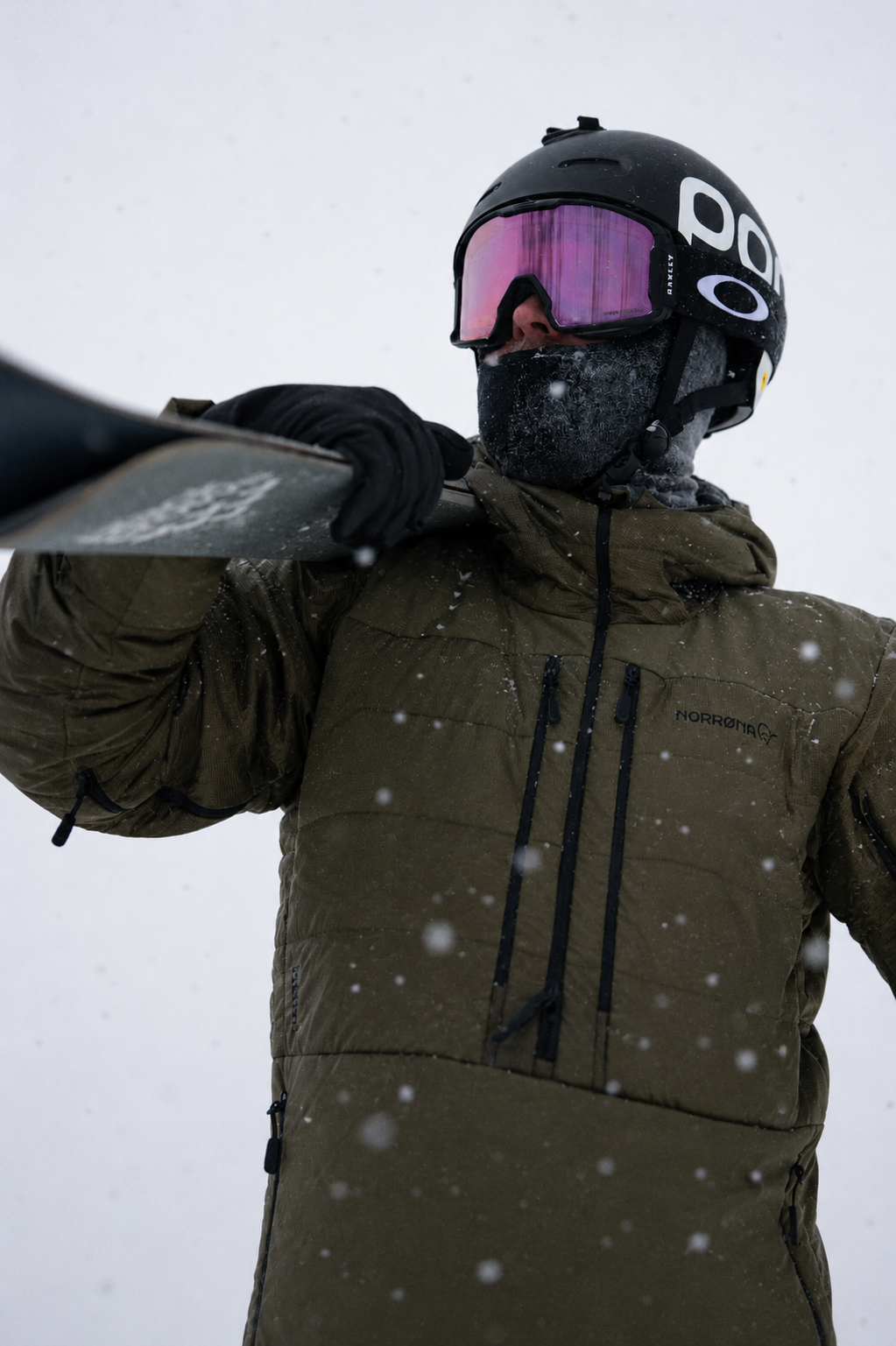 Skier wearing a Norrøna PrimaLoft®-insulated jacket, carrying a snowboard over their shoulder during heavy snowfall.