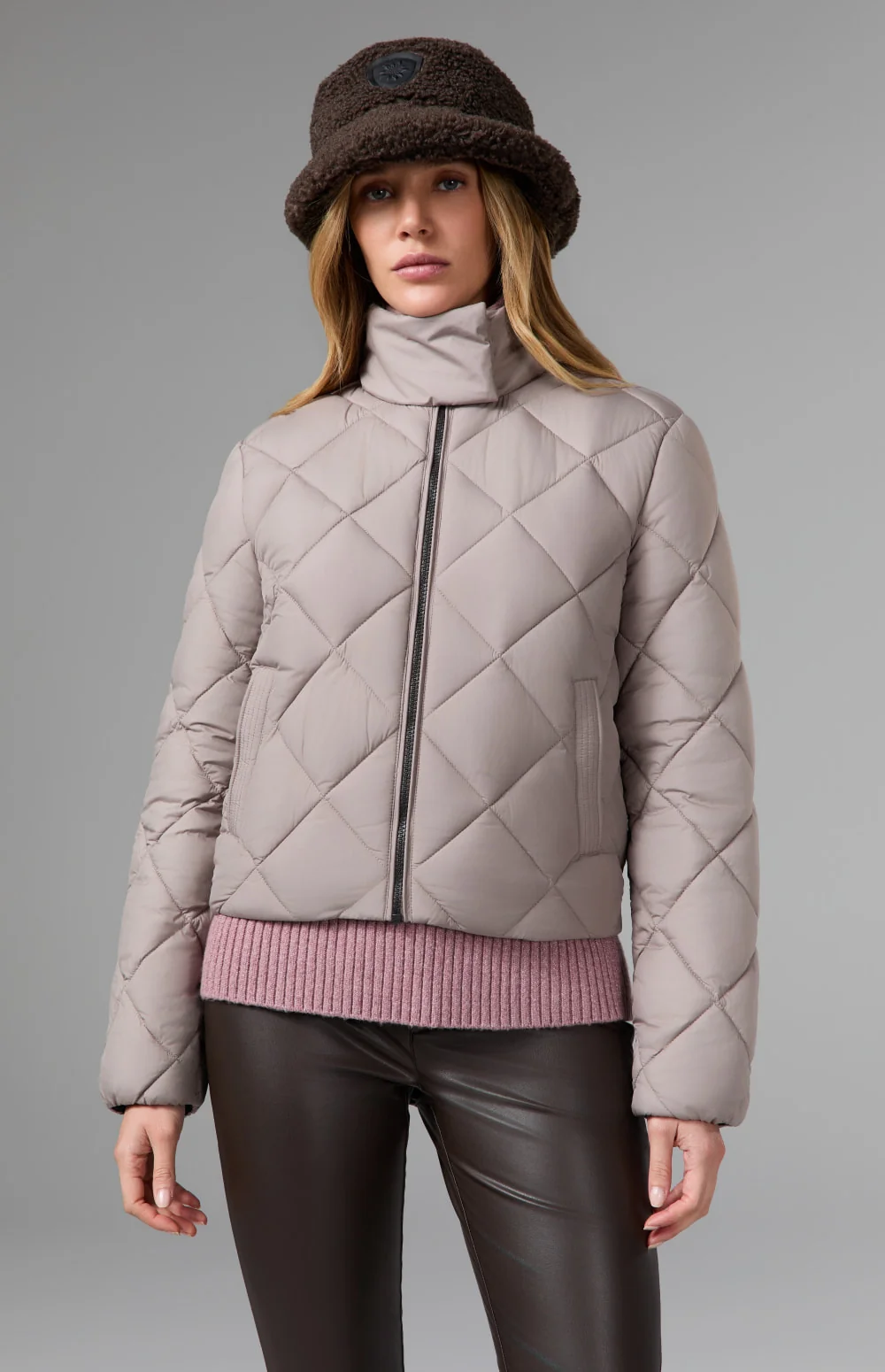 Woman wearing a taupe diamond-quilted funnel-neck jacket, fuzzy bucket hat, and brown leather pants.