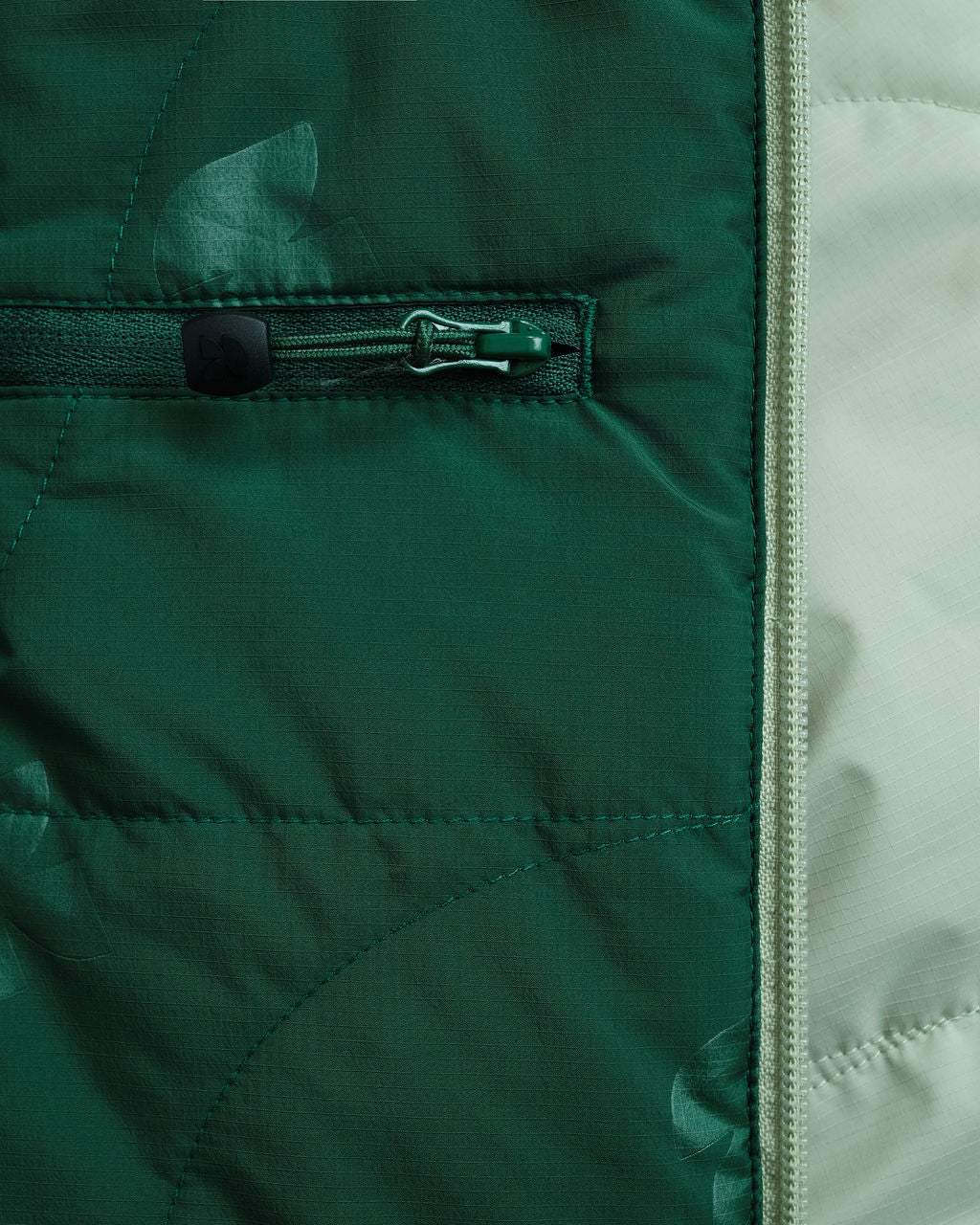 Close-up of the dark green printed lining of a jacket with a zippered pocket, beside the sage green outer fabric.