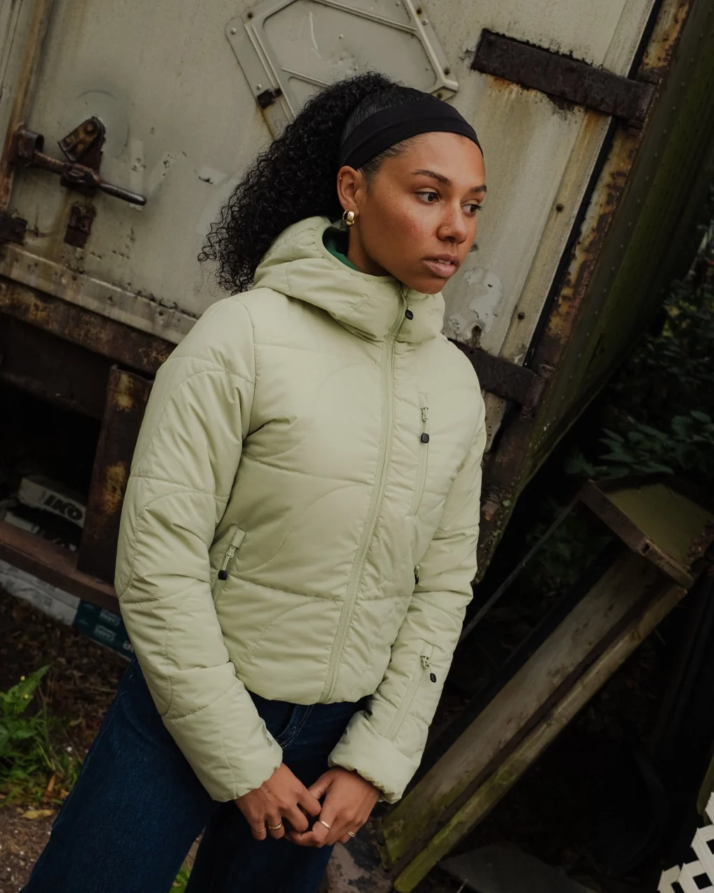 Woman wearing a sage green hooded quilted jacket and dark jeans, looking off to the side in front of a weathered metal trailer.