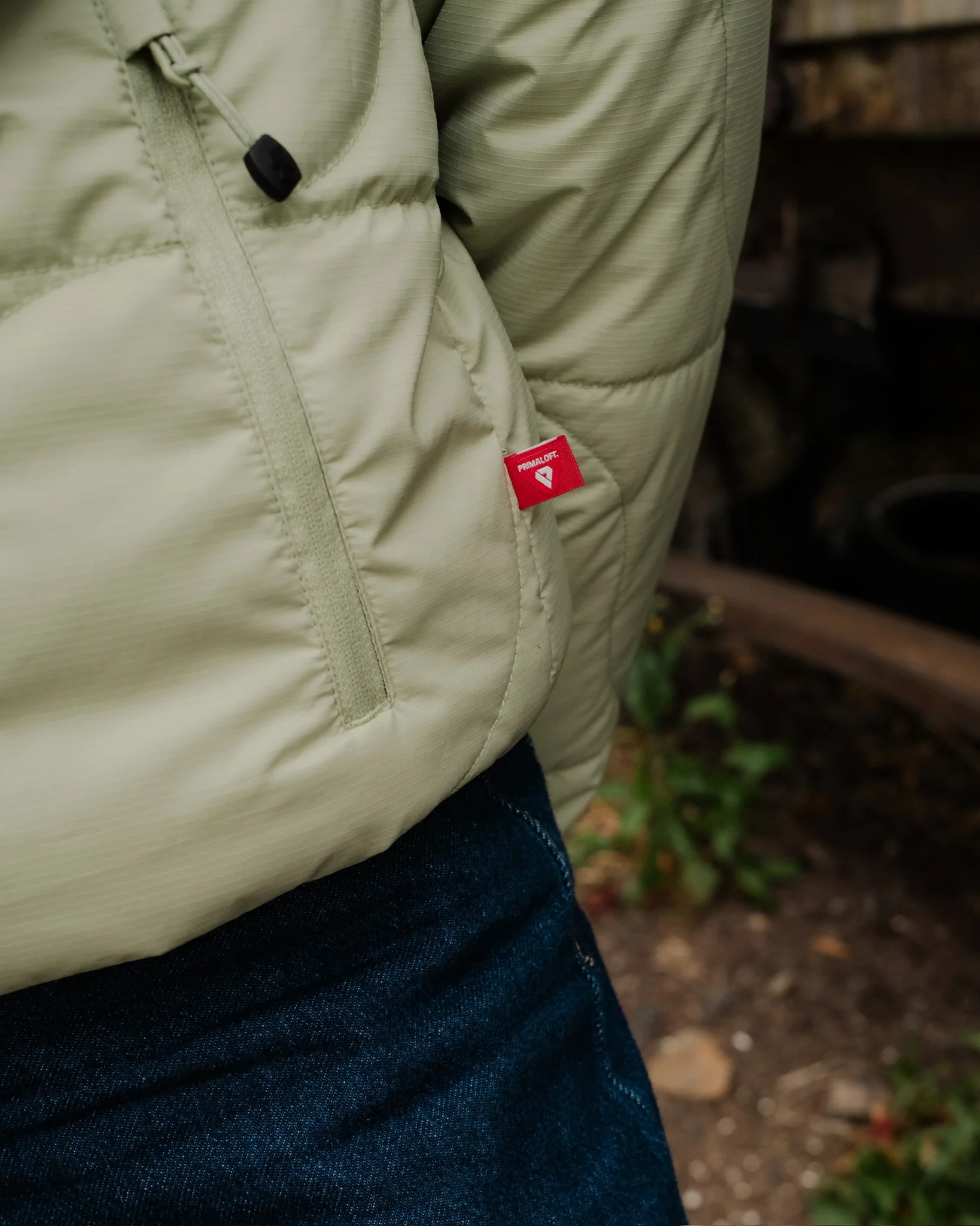 Close-up of the side of a sage green quilted jacket showing a zippered pocket, a black cord pull, and a red PrimaLoft® logo tag.