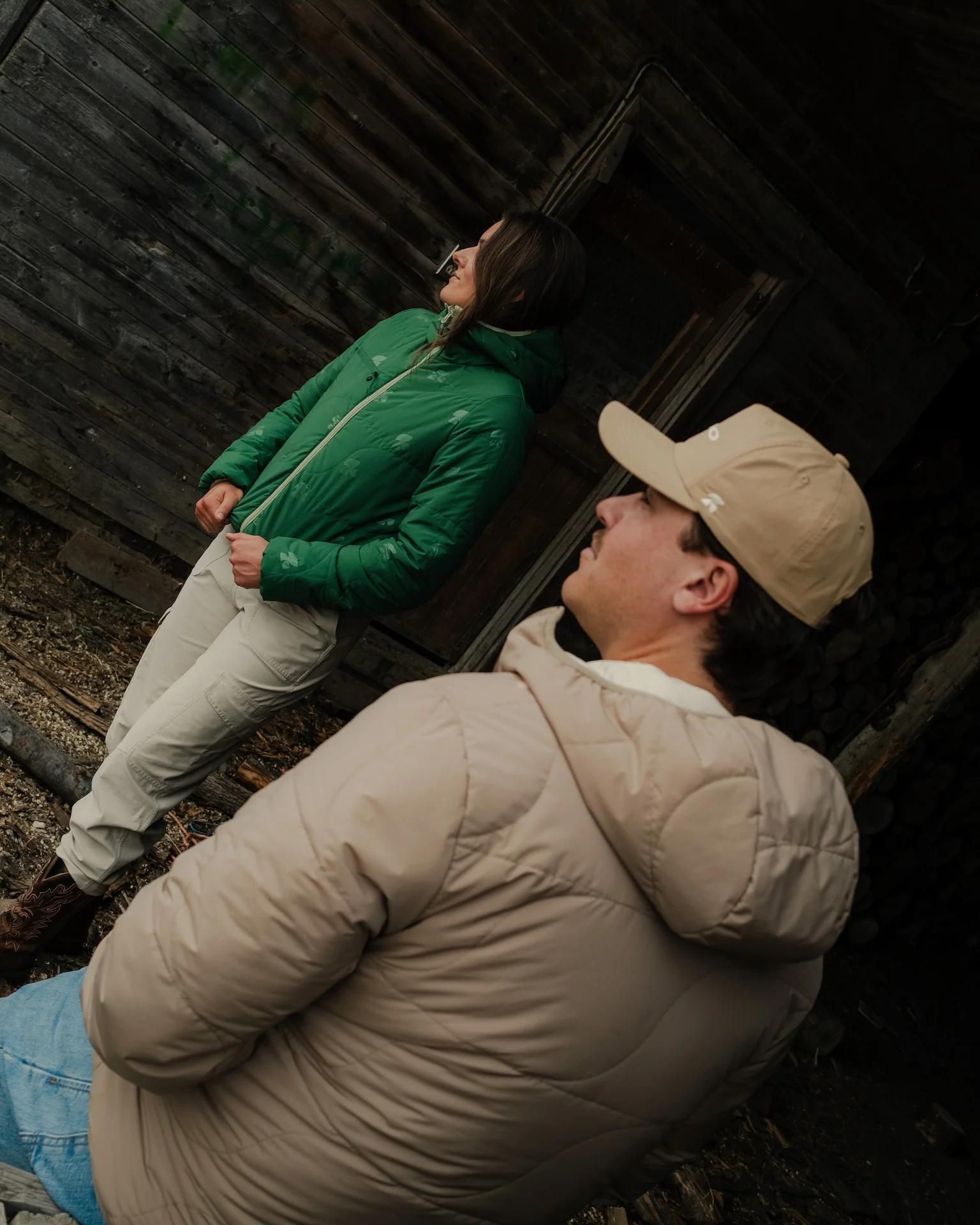 Woman in a green quilted jacket with logo print leaning against a dark wooden cabin, and a man in a tan cap and beige hooded jacket in the foreground.