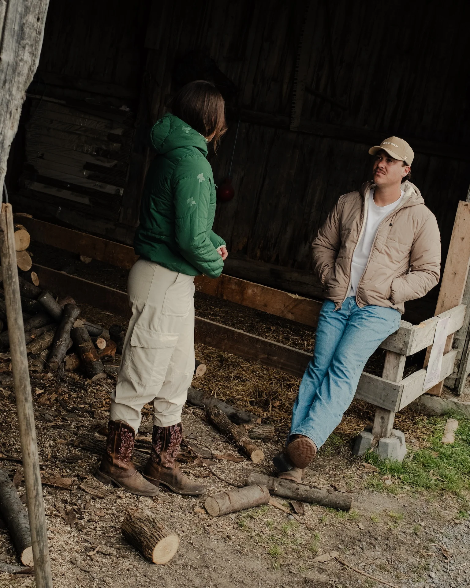 Woman in a green quilted jacket, cream cargo pants, and cowboy boots talking with a man in a tan cap, beige quilted jacket, and jeans sitting on a wooden fence in a barn.