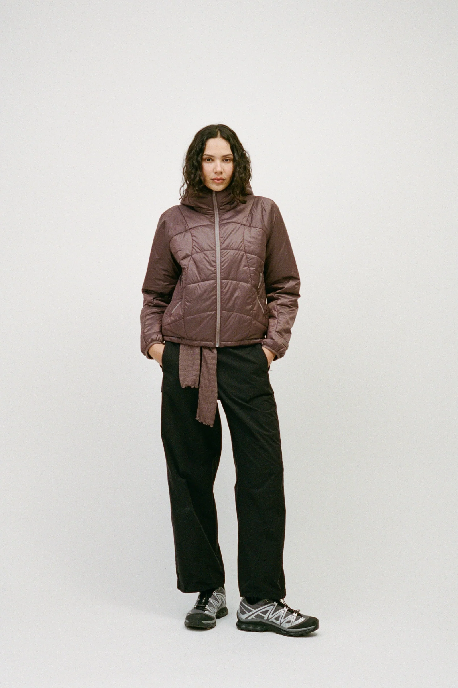 Woman wearing a mauve quilted puffer jacket with a sweater tied at the waist, black pants, and sneakers.
