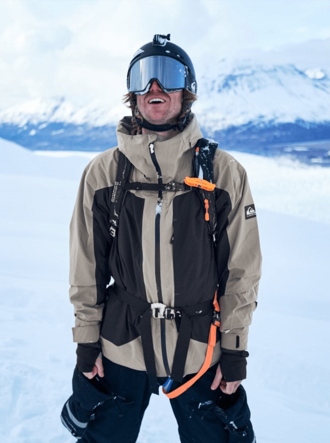 Person wearing a beige and black snow jacket, ski goggles, and a backpack, with snowy mountains behind.
