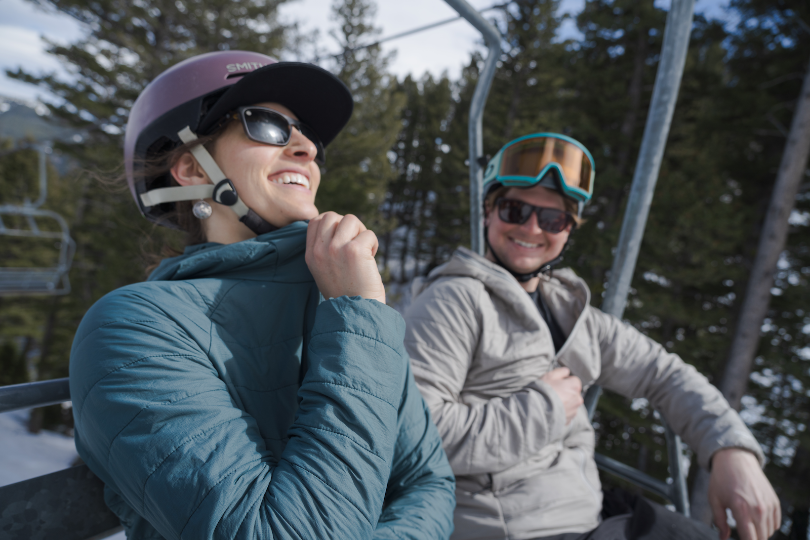 Two women sitting on a chairlift, smiling, wearing helmets and insulated jackets.