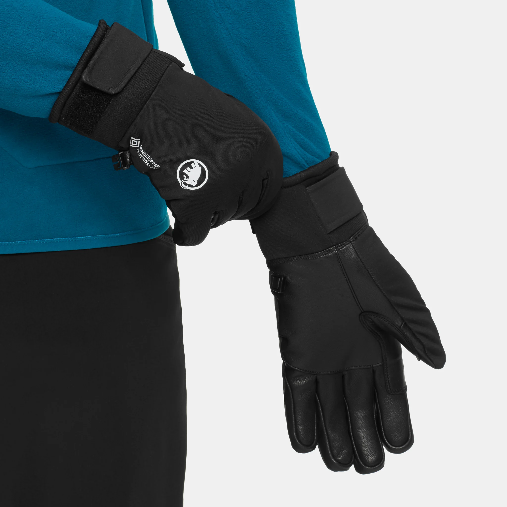 Close-up of a person in a teal long-sleeve top adjusting the wrist strap of a black Mammut glove.