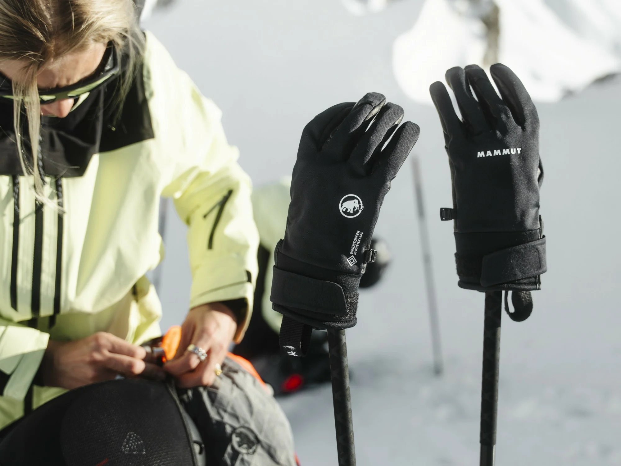 Black Mammut gloves hanging on ski poles while a woman in a pale green jacket works on a backpack.