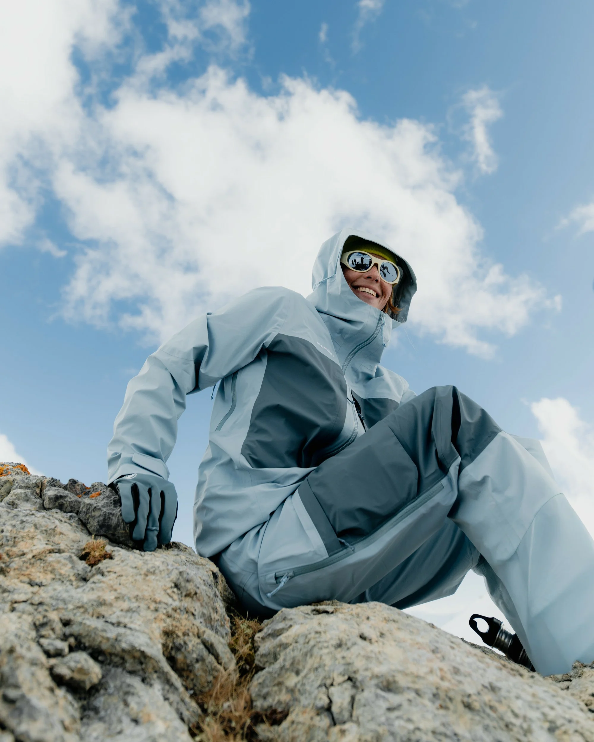 Person in a light blue ski suit and goggles sitting on a rock against a blue sky, low angle.