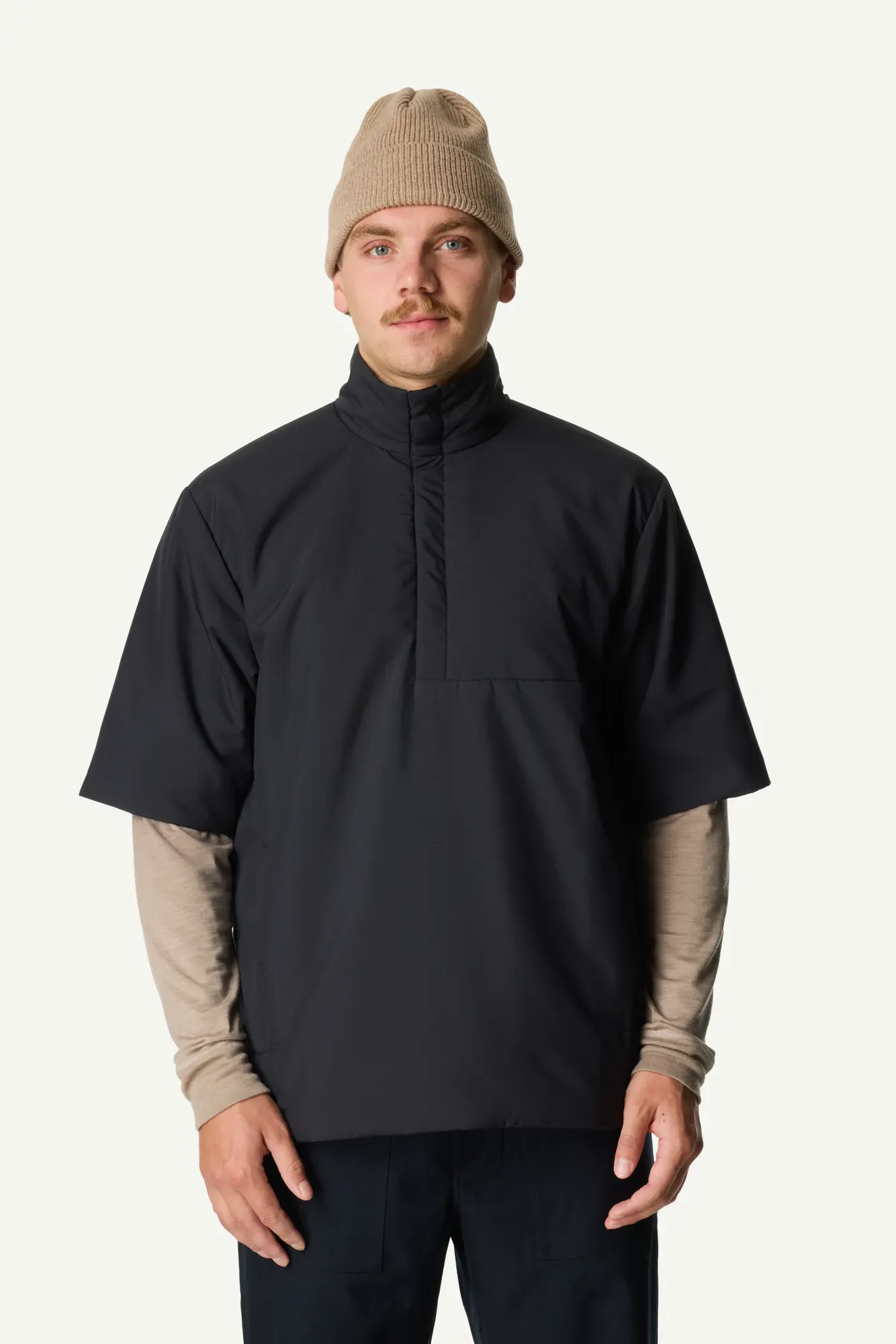 Man wearing a black short-sleeve high-neck pullover over a tan long-sleeve shirt and a beige beanie, front view.