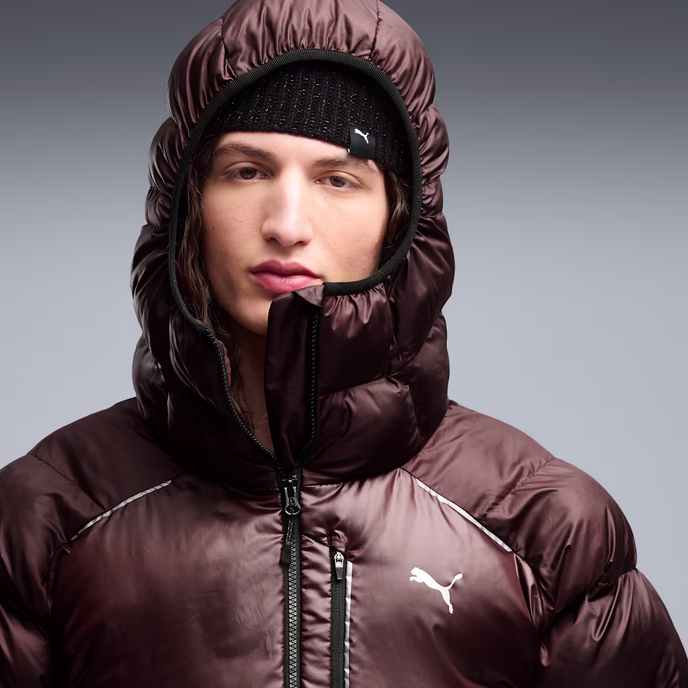 Close-up of a person in a brown hooded puffer jacket and black beanie, zipper open, Puma logo on the chest.