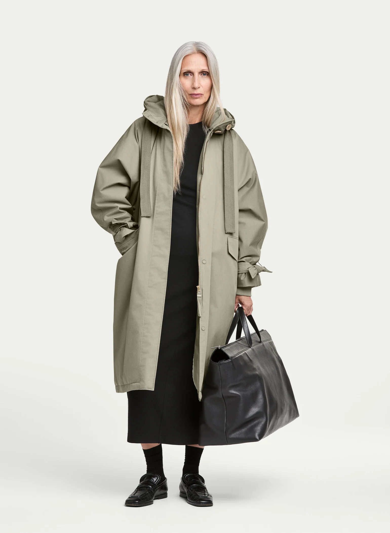 Woman wearing an oversized sage green hooded coat over a black dress, carrying a black leather tote bag.