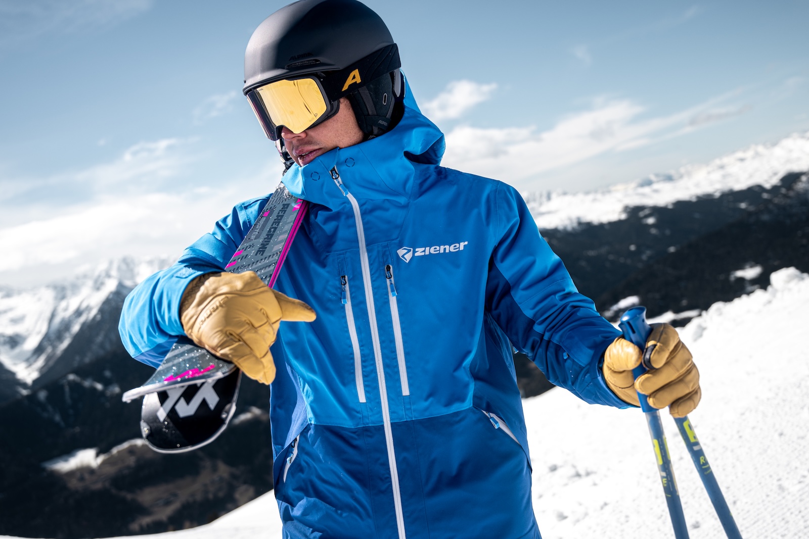 Skier wearing a blue Ziener jacket, helmet, and goggles, holding skis over one shoulder, mountains in the background.